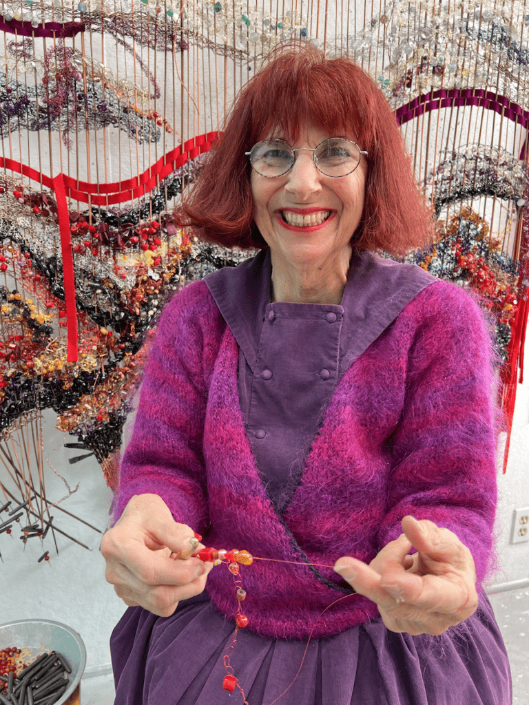 Artist Bonnie Meltzer holding beaded work in front of a piece included in the Burned Piano Project: Creating Music Amidst the Noise of Hate exhibition.